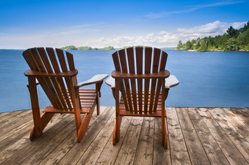 Wall Mural - Two Adirondack chairs sitting on a wooden dock facing a blue calm lake. Across the water is a green cabin nestled among green trees. 