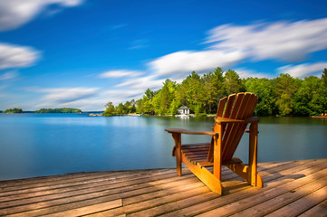 Wall Mural - Single Muskoka chair sitting on a wood dock facing a lake. Across the calm water is a white cottage nestled among green trees. There is a boat dock on the water in front of the cottage.