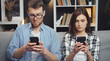 © MYDAYcontent - Spouses using their smartphones sitting on sofa next to each other consumed with own stuff, front view
