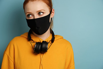 A girl in a yellow hoodie and a black protective mask with headphones around neck stands on a blue background, looks to the side