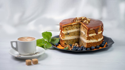 Cup of coffee with two lumps of sugar on a saucer and next to a cut carrot cake, covered with caramel and decorated with nuts on top, behind a white textile background