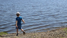 A Boy In Blue Clothes From The Back Walks Along The Rocky Shore Of A Lake Or River To The Small Waves Rolling On The Stones