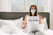 © LIGHTFIELD STUDIOS - female freelancer in medical mask holding Stay at home sign and working on laptop during self isolation in bed