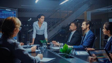 Wall Mural - Late at Night in Meeting Room: Confident Female Executive Director Stands in the Head of the Conference Table Leans on it and Delivers Ground Breaking Speech to a Group of Businesspeople and Investors