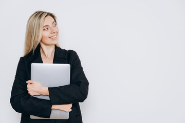 A pretty young smiling woman is standing with a notebook pc under her arm against a light background