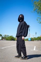 Wall Mural - Young man with orange dyed hair in a black hoodie with trousers and sports boots in a protective mask and black sunglasses against a blue sky. Urban view.