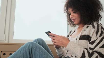 Wall Mural - Young pleased positive african curly woman indoors at home using mobile phone