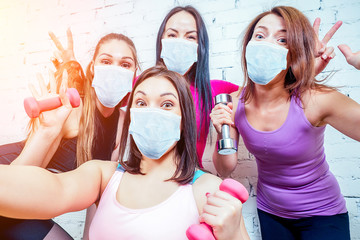 four beautiful and young women girlfriends in medical mask are photographed selfie on the phone in sportswear in the gym. group portrait of a woman self having fun with dumbbells