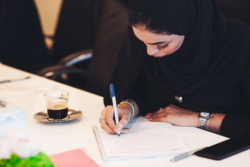 Pensive Arab woman planning working schedule writing in notebook while sitting at working place, Arabic female administrative manager making notes of information browsed on netbook.