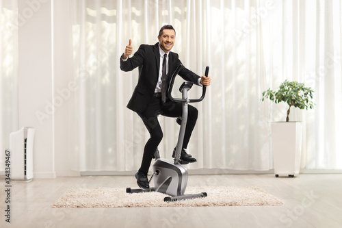 Cuadros en Lienzo Businessman riding an exercise bike at home and showing thumbs up