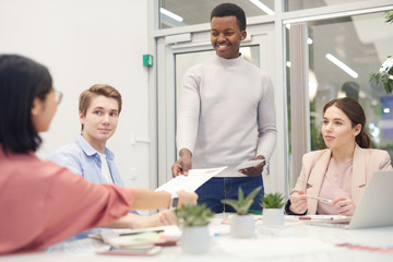 Wall Mural - Multi-ethnic group of young business people collaborating on work project during meeting in office, focus on smiling African-American man handing documents