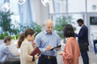 © Seventyfour - Group of three business people talking while standing in modern office interior, focus on bald adult businessman talking to two women