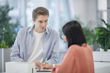 Waist up portrait of young man talking to Asian friend while standing at tall counter in university, copy space