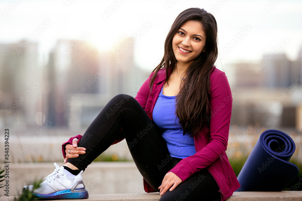 Portrait of a beautiful mixed ethnicity brunette woman in sportswear, sitting relaxed after a morning yoga workout with city skyline in the background - obrazy, fototapety, plakaty 