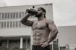 young strong athlete male drinking water from black bottle during outdoor fitness workout on university building backgroung