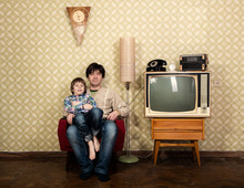Little Boy With Father Sitting On Armchair In Room, Interior From 70s 20th Century, Retro Stylization, Image Toned. Family At Home With Old Tv Set And Rarity Appliances And Furniture
