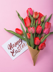 Wall Mural - red  beautiful tulips in an ice cream waffle cone with card Happy Easter on a color background. Conceptual idea of a flower gift. Spring mood