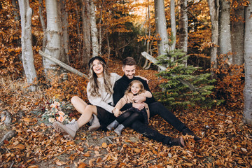 Wall Mural - Stylish family in the autumn forest. A young guy and a girl are sitting on yellow leaves near a wooden fence with their daughter.