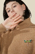 A brunette lady with a blunt bob haircut is covering her mouth. The lady's beige sweater is decorated with an elegant brooch made as a golden dragonfly with malachite enamel and rhinestones. 
