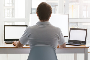 A man is sitting at a workplace with two laptops and a monitor near the window. Remote work from home in quarantine. Back view