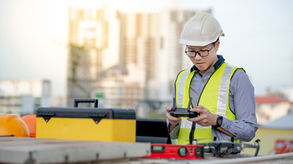 Wall Mural - Asian engineer man working with drone, laptop and working tools at construction site. Male worker using unmanned aerial vehicle (UAV) for land and building site survey in civil engineering project.