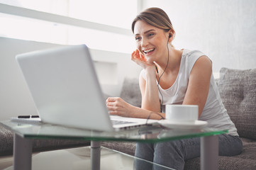 Attractive happy young student studying online at home, using laptop computer, headphones, having video chat, waving. Remote work, distance education. Video conference or virtual event on quarantine