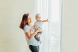 © Юлия Завалишина - Stay Home Stay Safe. Young mother holding her child. Woman and infant little girl relaxing in white bedroom near windiow indoors. Happy family at home. Young mom playing whith her daughter.