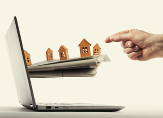 Newspaper with paper houses stick out of a laptop screen. The human buying a house via internet. Concept of modern technologies in real estate industry.