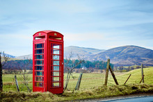 Bright Red Iconic British Telephone Box. This Old Fashioned Phone Booth Is In A Rural Landscape Beside Country Lane, With Fields And Scottish Hills In The Background.