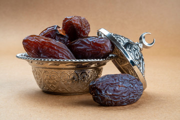 Religious iftar Ramadan concept: Side view of few dried organic sweet date fruits in a silver traditional bowl with ornate cap on brown background for Muslim fasting break.
