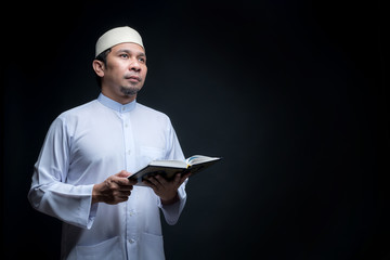 Portrait Asian Muslim man standing and reading the Quran and appreciates on black background, with copy space.