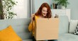 © VAKSMANV - Young pretty woman sitting on couch in living room and opening carton box. Cheerful girl getting parcel at home. Mail delivery to house.