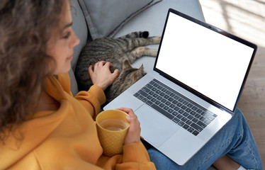 Wall Mural - Young hispanic latin teen girl student relax sit on sofa with cat holding laptop looking at mock up white computer screen online learning on pc, elearning, watching movie. Over shoulder closeup view