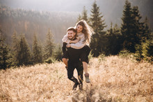 Stylish Model Couple In The Autumn Mountains. A Young Guy And A Girl Run Along The Slope Against The Background Of The Forest And Mountain Peaks At Sunset.