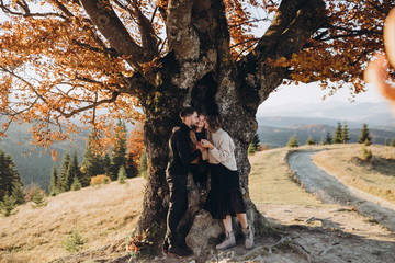 Wall Mural - Stylish young family in the autumn mountains. A guy and a girl with their daughter stand under a large old tree against the background of a forest and mountain peaks at sunset