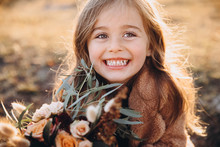 Portrait Of A Small Stylish Beautiful Model Girl Who Stands In The Autumn Mountains At Sunset And Holds A Bouquet Of Flowers In Her Hands