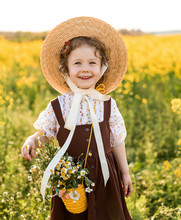 Portrait Of Smiling Cute A Lilttle Girl With Curly Hair And A Straw Hat, Stylishly Dress,  Bag Over Her Shoulder Full Of Daisies Among Of A Flowering Field.
