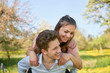 © stock mp - Young couple in nature having fun, a boyfriend carrying his girlfriend on his back on a meadow with yellow flowers and trees, in the background a bicycle and blue sky