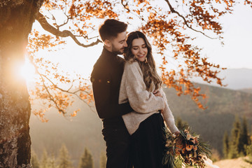 Wall Mural - Stylish young couple in the autumn mountains. A guy and a girl stand together under a big old tree against the background of a forest and mountain peaks at sunset. The girl has a bouquet in her hands