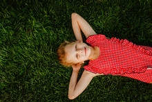 Young Blond Cute Girl In Red Squared Summer Swimsuit Laying On Green Grass