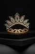 Subject shot of a tiara made as golden leaves adorned with emerald green gems and clear sparkling rhinestones. The luxury queen crown is fixed on the black stepped surface.