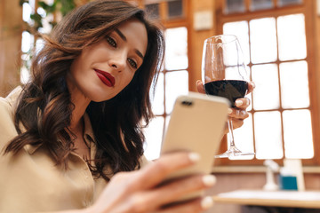 Wall Mural - Image of pleased adult woman drinking red wine and using mobile phone