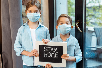 sister and brother in medical masks holding chalkboard with stay home lettering