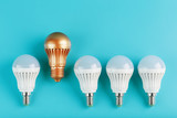 Fototapeta  - One gold led light bulb is higher and stands out from a row of white lamps on a blue background.