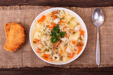 Homemade noodle soup with pieces of chicken breast, carrots, potatoes and parsley in a white plate and two slices of toast, top view