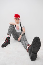 Portrait Of A Cool Fashionable Modern Young Girl. A Short Haircut With Shaved Temple. Dyed Bright Red Hair. Red Lipstick. Studio Photo On White Background. Suspenders And Checkered Pants.