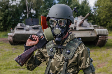 A soldier in a gas mask with a Kalashnikov rifle against the background of armored vehicles.