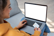 Happy Hispanic Female Shopper Customer Holding Credit Card Paying Online On Laptop Mock Up White Screen Shopping In Web Store Making E Commerce Secure Digital Payment On Website. Over Shoulder View.