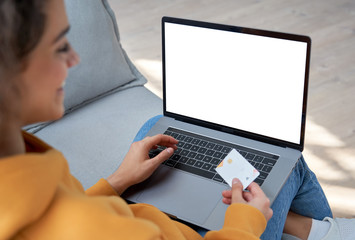 Happy hispanic female shopper customer holding credit card paying online on laptop mock up white screen shopping in web store making e commerce secure digital payment on website. Over shoulder view.