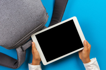 Wall Mural - Kid hands with digital tablet computer and gray backpack over light blue background. Top view